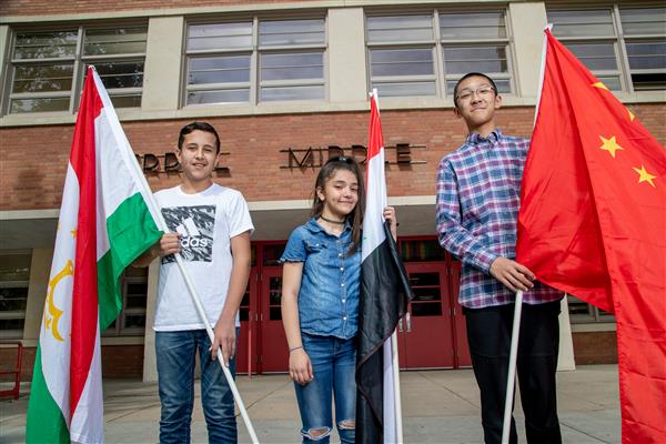 Three students stand in front of their school holding flags from other countries  